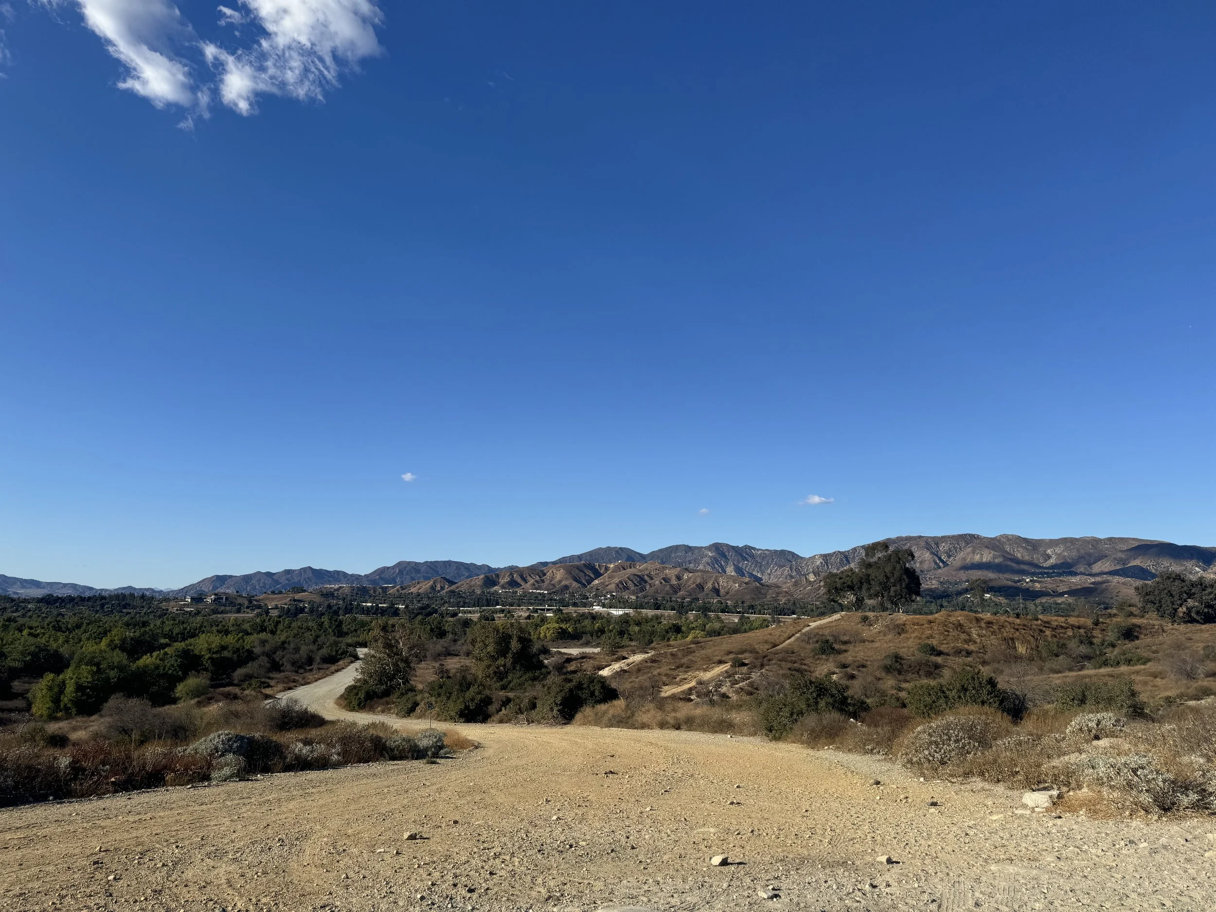 A clear blue sky over a rugged dirt road winding through hilly terrain with sparse vegetation and distant mountains.