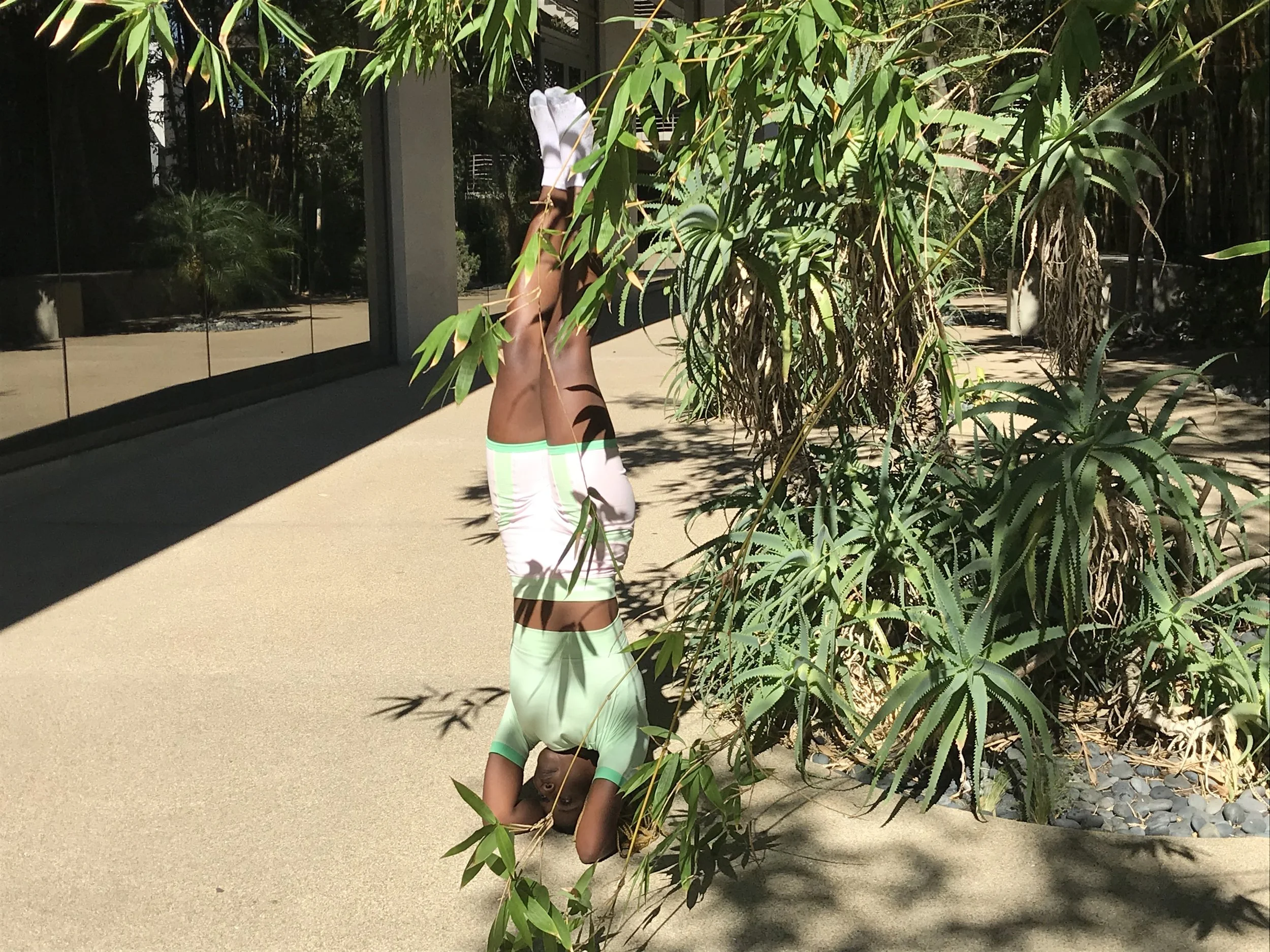 A woman in a green shirt and white shorts performing a headstand outdoors on a paved surface, with leafy plants and a modern building reflected in windows nearby.