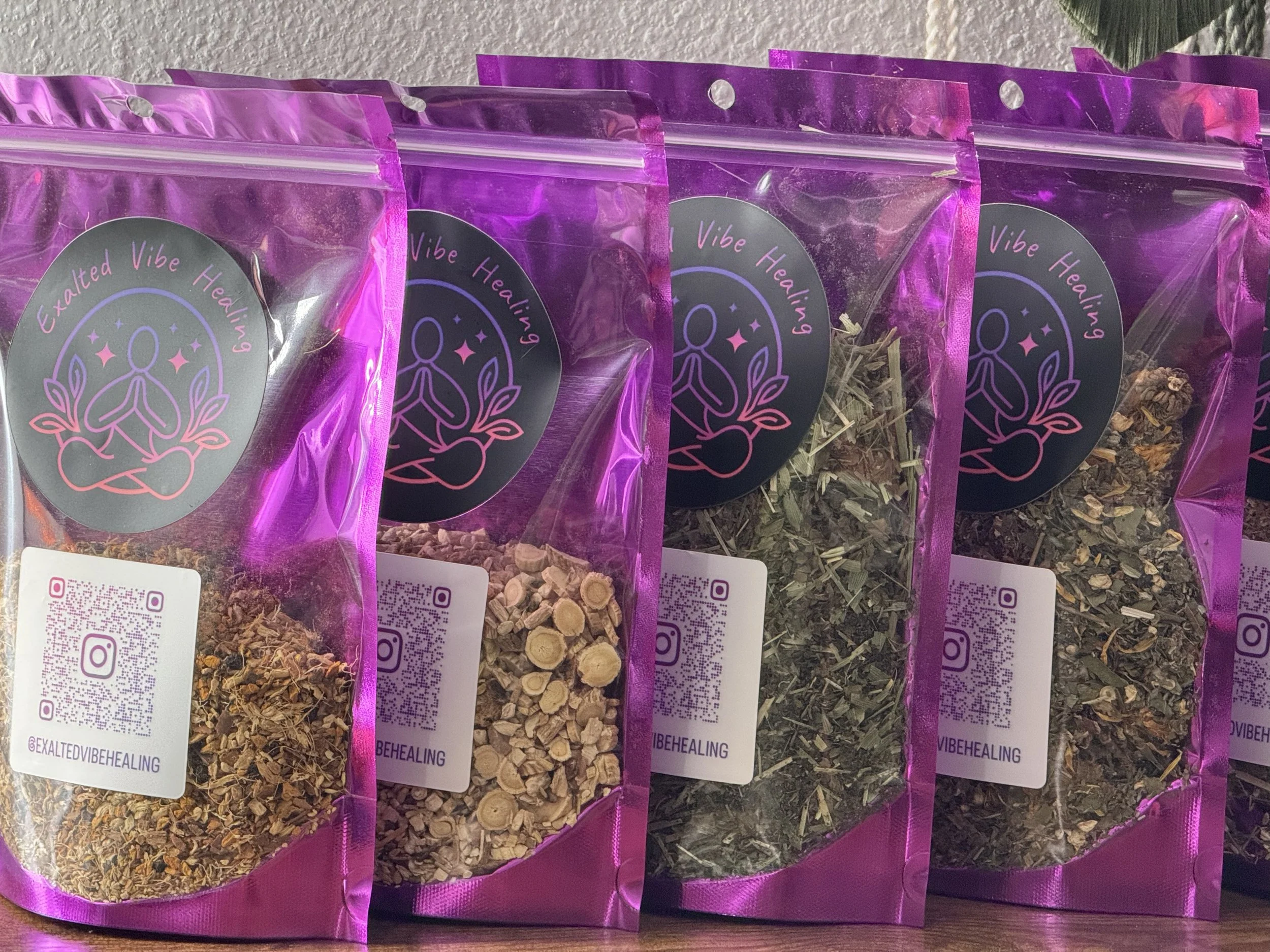 Four purplish pink resealable bags of dried herbal or botanical products, each with a black circular logo featuring a stylized person meditating, surrounded by leaves and stars, and labeled "Exalted Vibe Healing" with a QR code and Instagram handle.