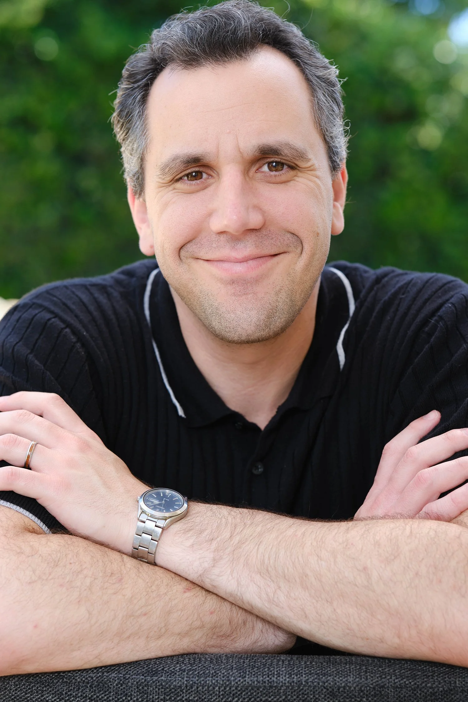 Jackson Kaplan - Author headshot. A man with brown and gray hair, wearing a black shirt, with his arms crossed on a chair, a silver watch on his left wrist, set outdoors with a blurred green natural background.