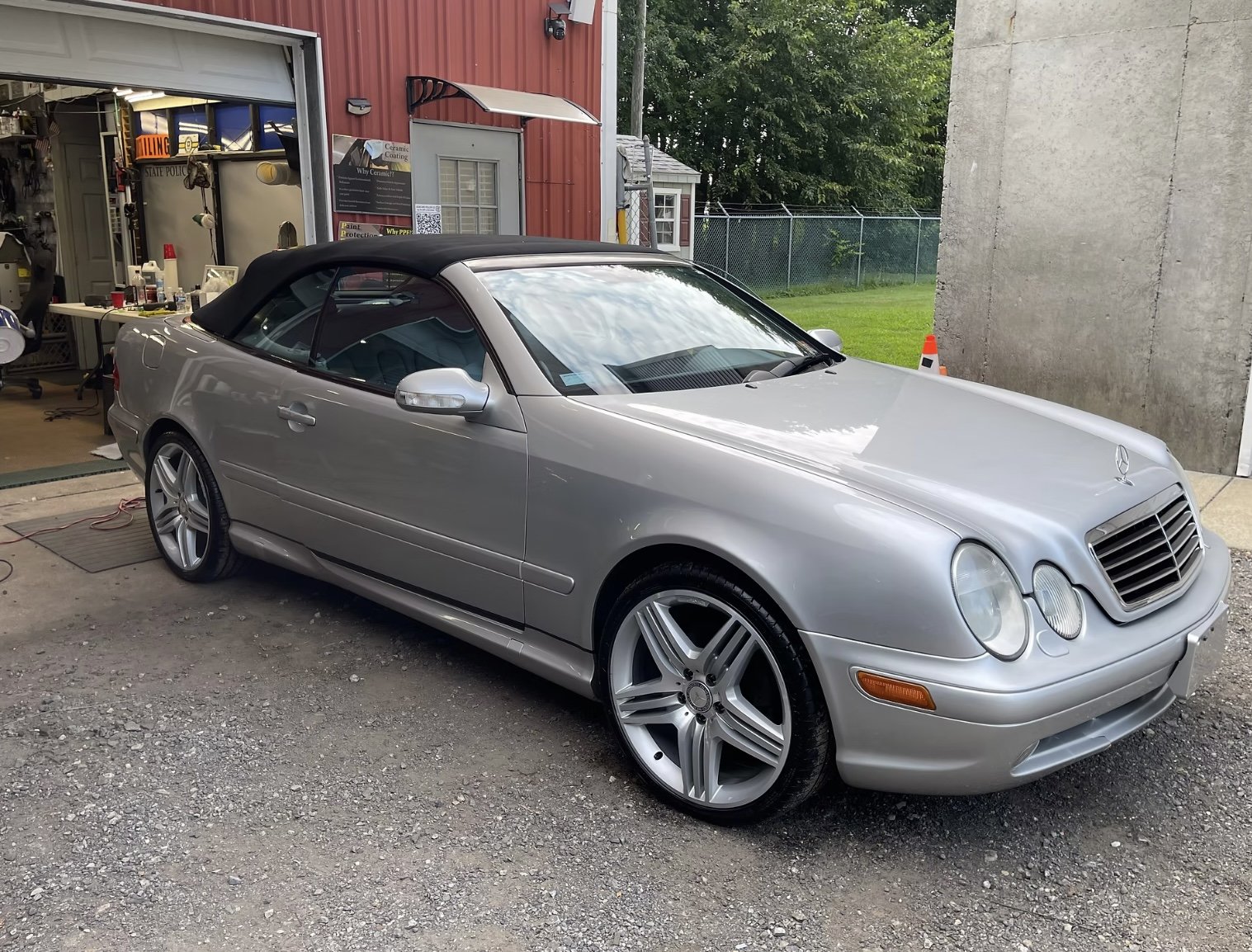 A silver Mercedes-Benz convertible parked outside a garage with a black soft top roof. The garage contains tools and supplies, and there is a red building with signs on the wall behind the car. The setting is outdoors with green grass and trees in th