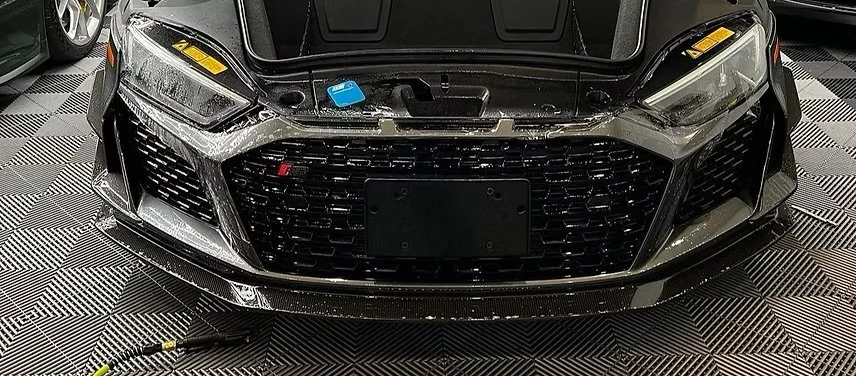 Close-up of a black car's front end, showing the grille, headlights, and front bumper in an indoor setting with a checkered floor.