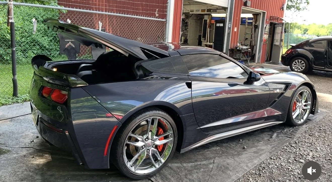 A black sports car with open rear hatch, parked outside a garage. The car features sleek, aerodynamic design with shiny rims and red brake calipers. The background includes a chain-link fence, a red building, and another vehicle.
