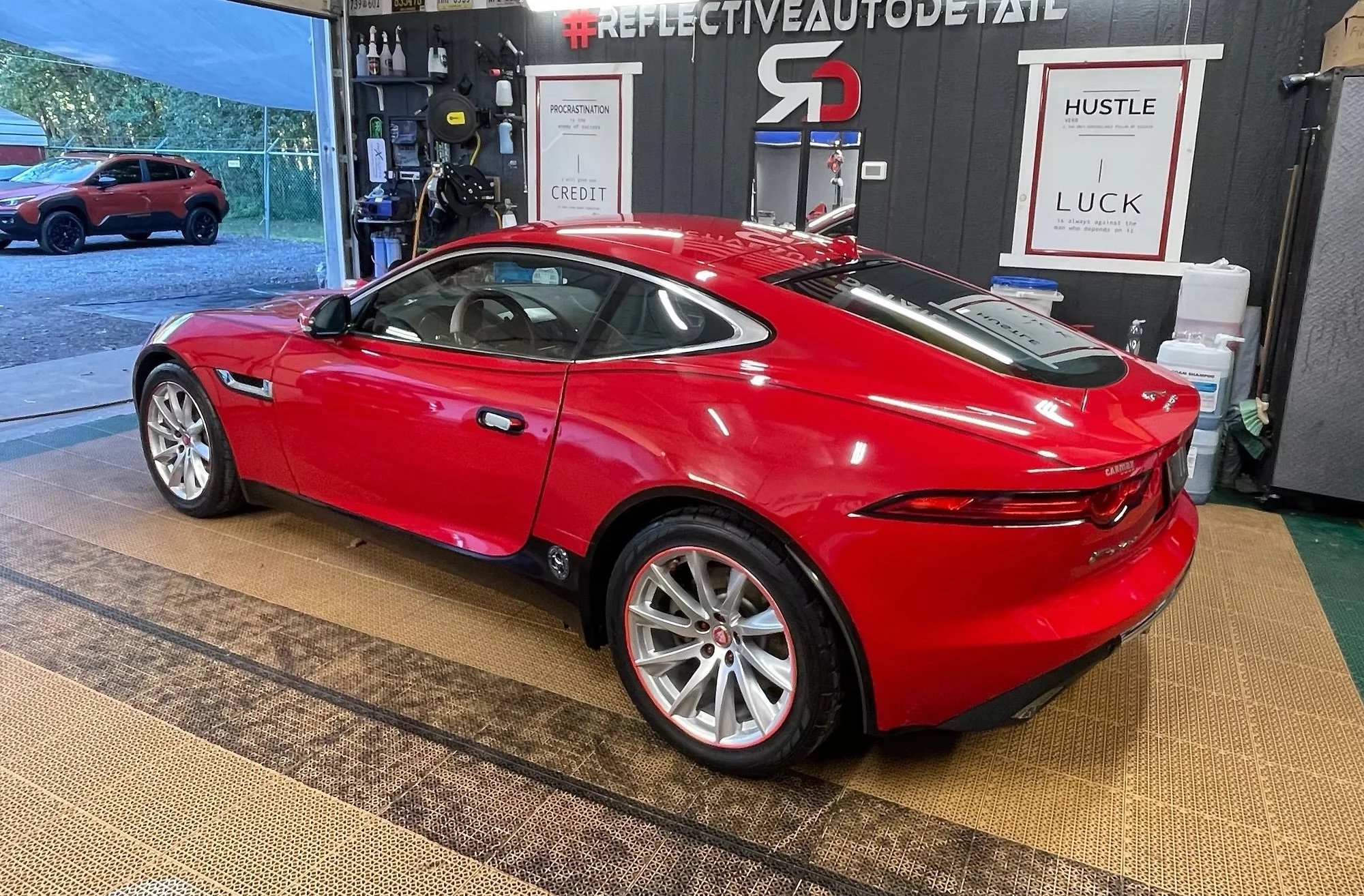 A red Jaguar parked inside a garage with a dark wall. Through the garage door, an outdoor area with a red vehicle and a blue canopy can be seen. The garage wall has motivational posters and tools organized on shelves.
