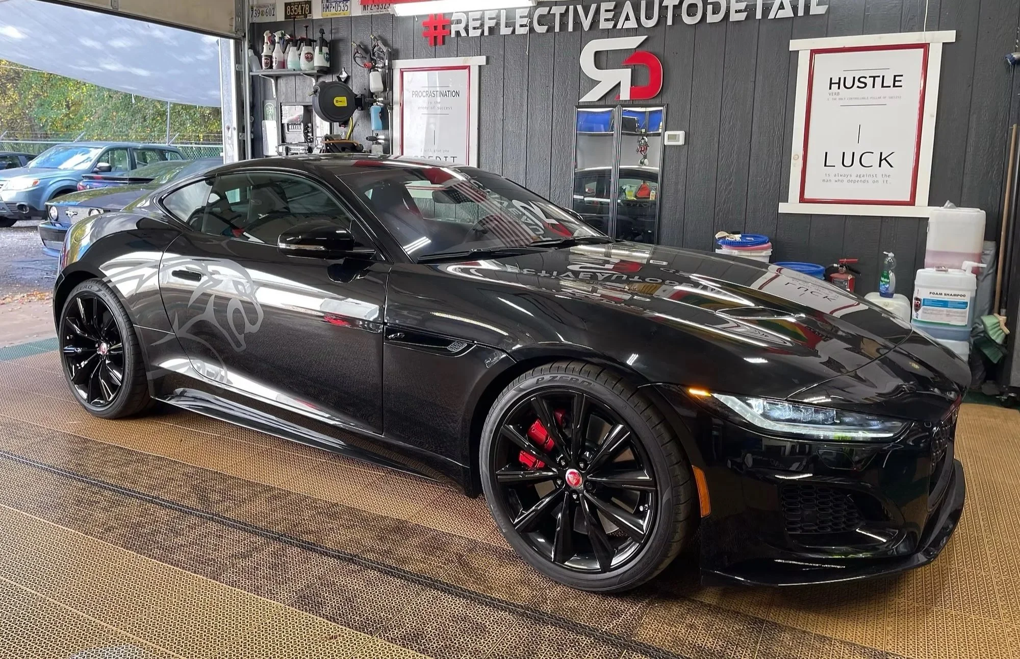 A black sports car parked inside a garage with a sign that says '#REFLECTIVEAUTODETAIL'. The garage has signs with motivational quotes and various car cleaning supplies and equipment. Outside the garage, several parked cars are visible.