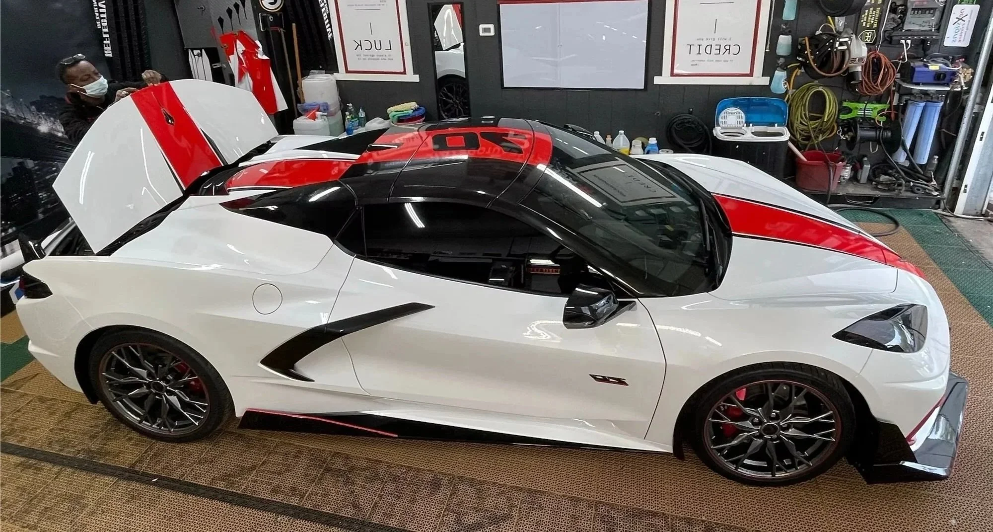 A white sports car with black and red accents parked inside a garage with tools and equipment on the walls.