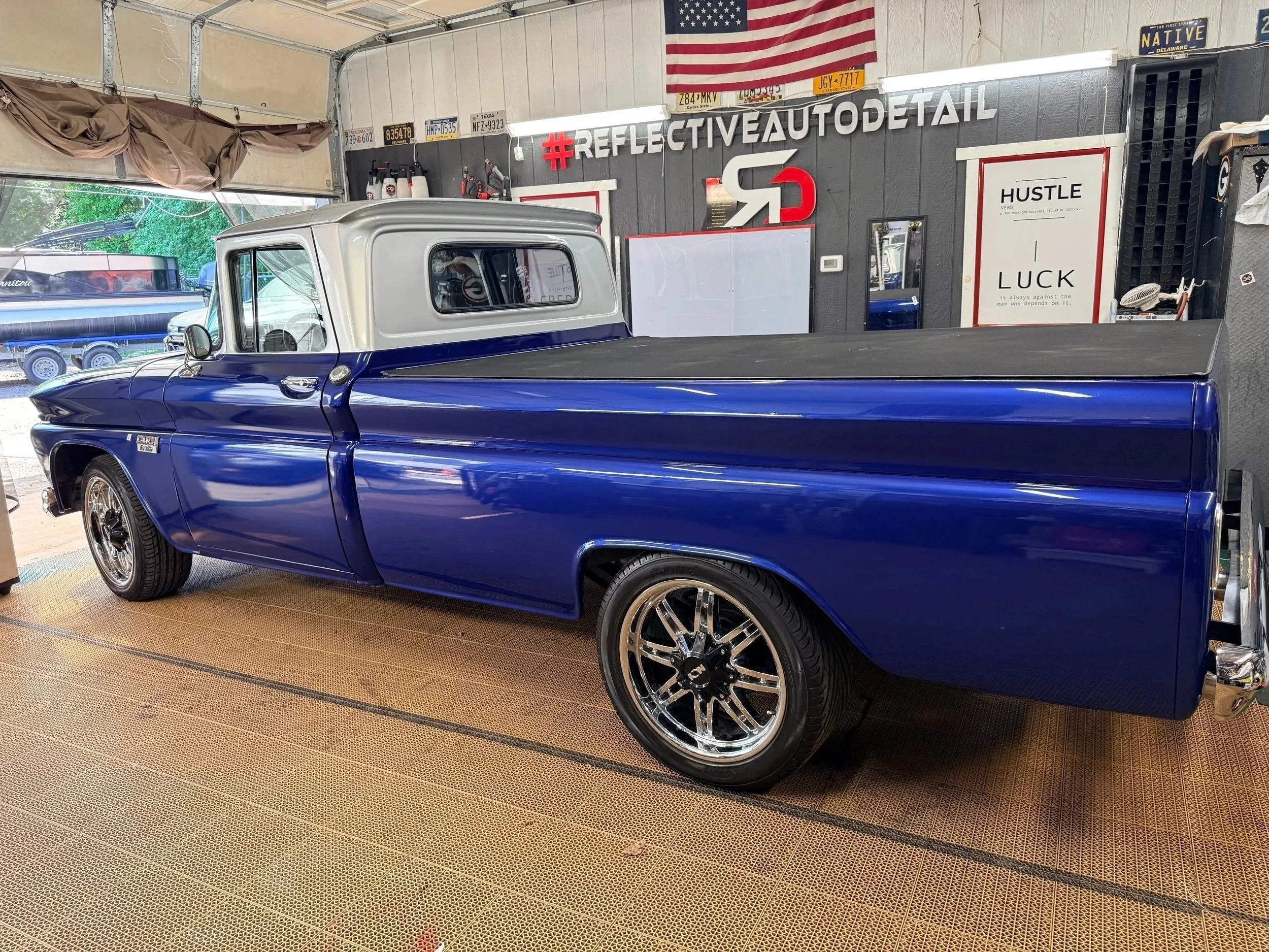 A classic blue pickup truck with a white top parked inside a garage. The garage wall features patriotic flags, license plates, and automotive signs.