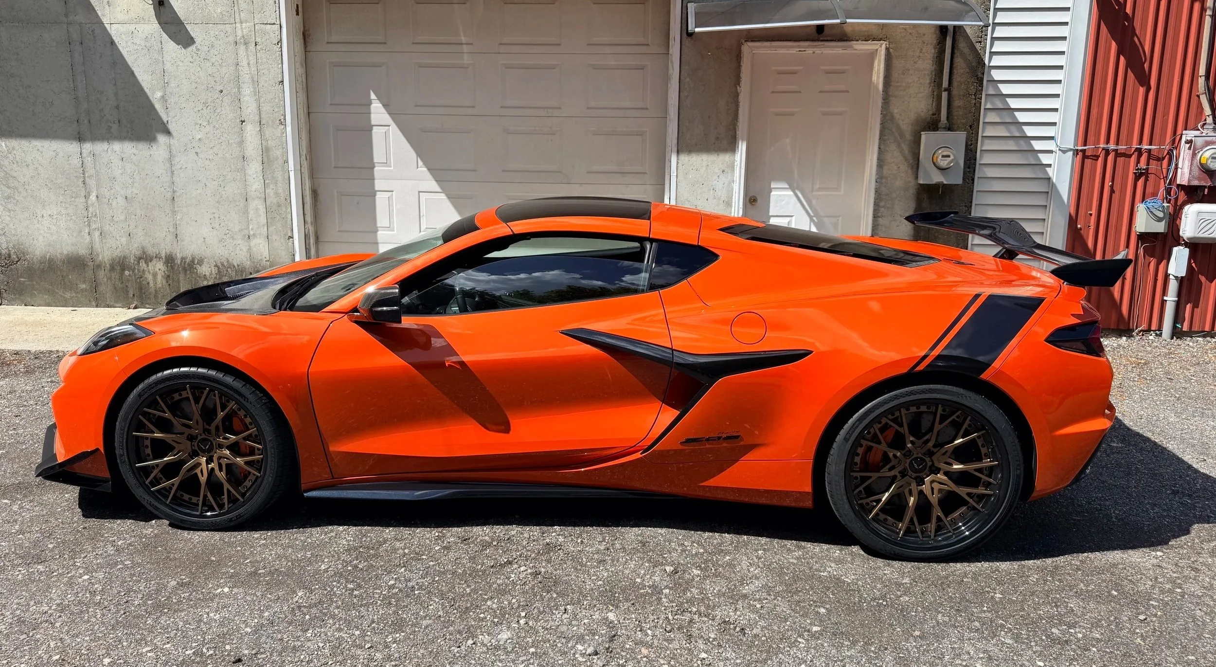 An orange sports car with black accents parked in front of a beige garage door and a red building with white siding.