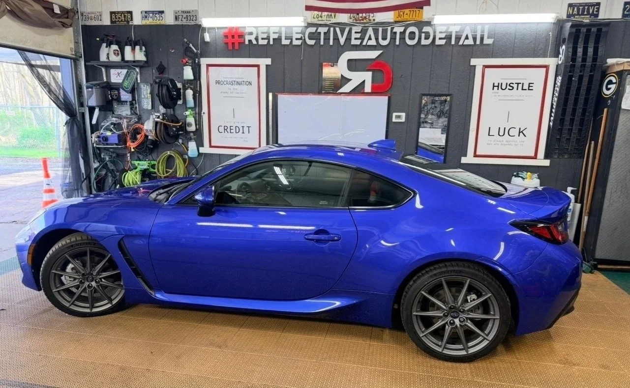 A blue sports car parked inside an auto detailing garage with tools and supplies on the wall behind it. The wall features motivational posters and a hashtag sign reading '#REFLECTIVEAUTODETAIL'.