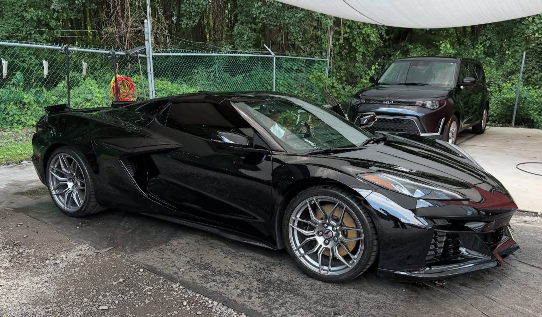 Black sports car parked outdoors next to a curb, with a dark SUV parked nearby and a chain-link fence with greenery in the background.