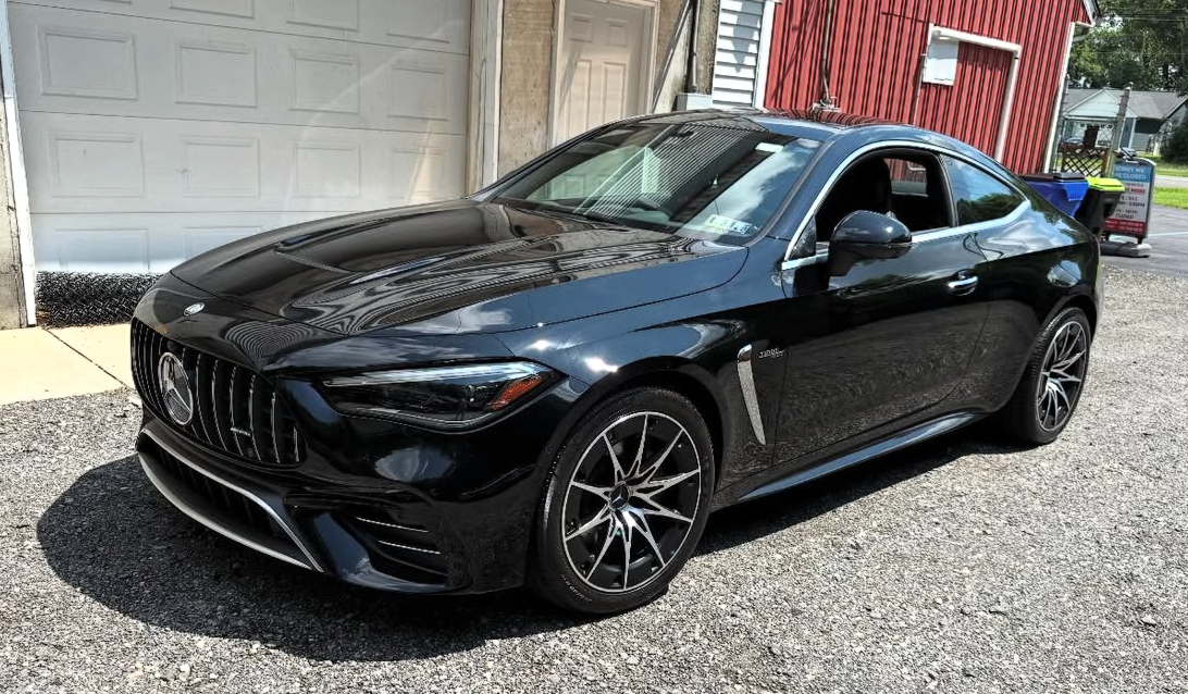 Black Mercedes-Benz AMG sports coupe parked on a gravel driveway in front of a garage and a red building.