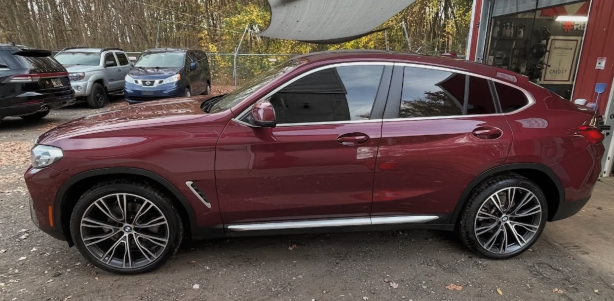 A red BMW SUV parked outside a garage with several other vehicles in the background and autumn trees.
