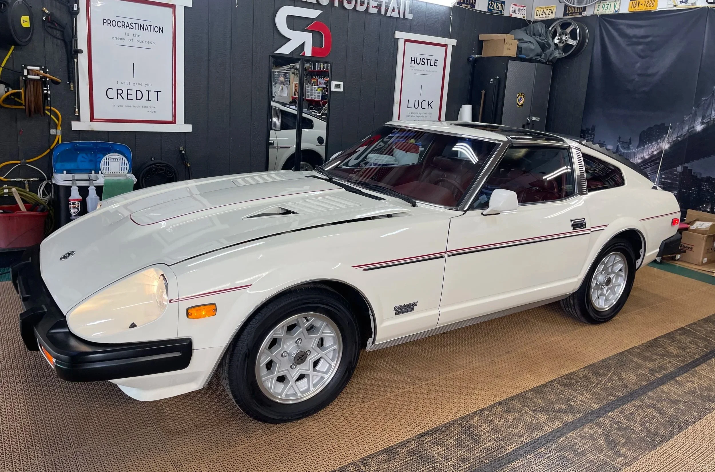 A white vintage car with pop-up headlights parked inside a garage or detailing shop. The background has signs with motivational quotes, license plates, a flag, and tools hanging on the wall.