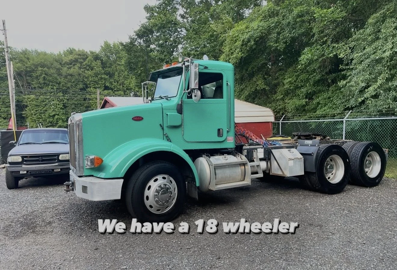 A teal semi-truck with six wheels parked on gravel ground, with a gray car behind it and a green fence with trees in the background. Text on the image reads, "We have a 18 wheeler."