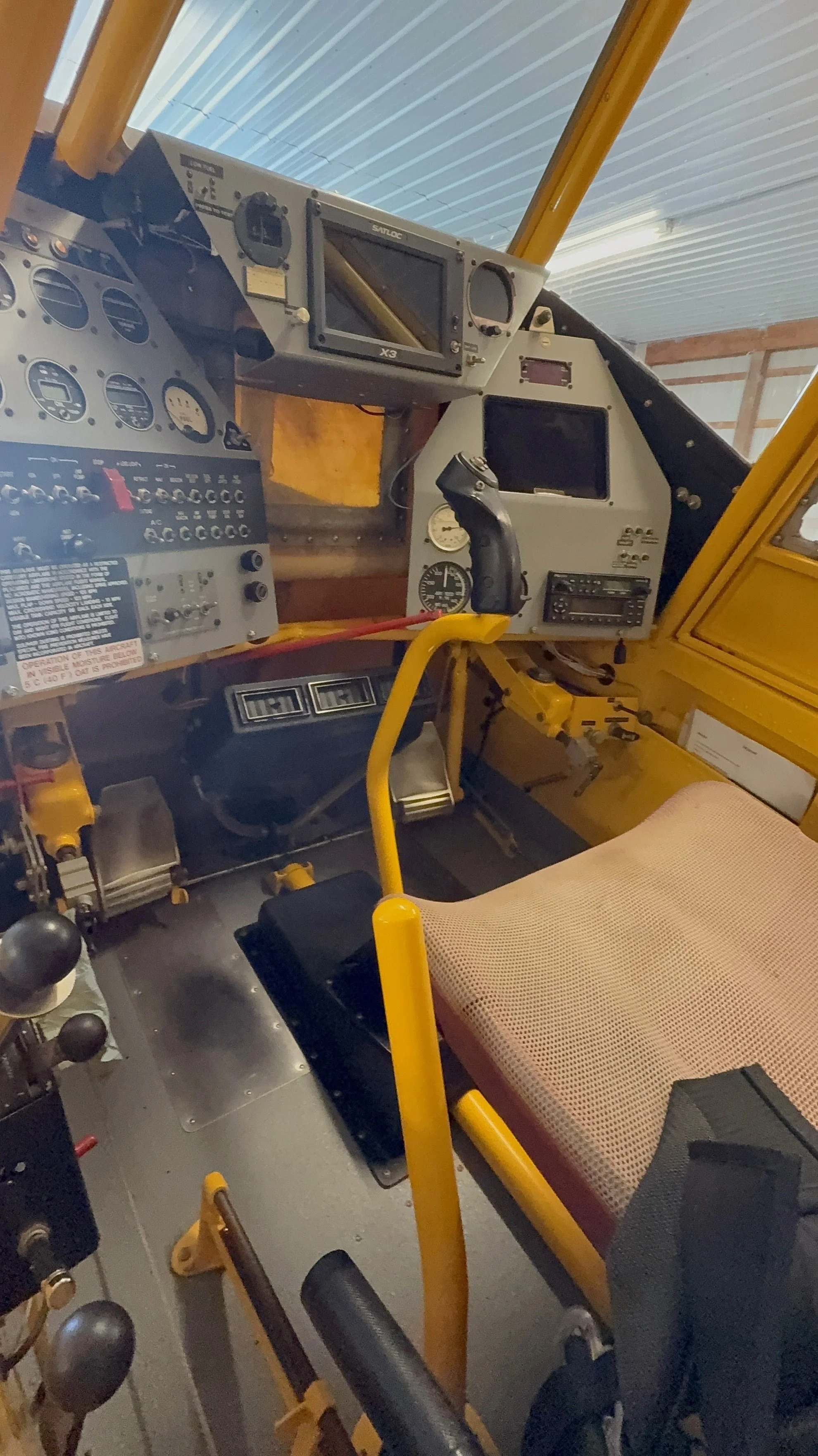 Interior of an aircraft cockpit showing various instruments, gauges, control sticks, and a beige seat with a yellow frame.