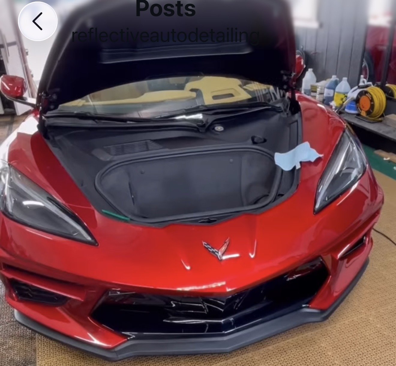 Red Chevrolet Corvette sports car with the hood open in a garage or workshop, with tools and bottles on a workbench in the background.