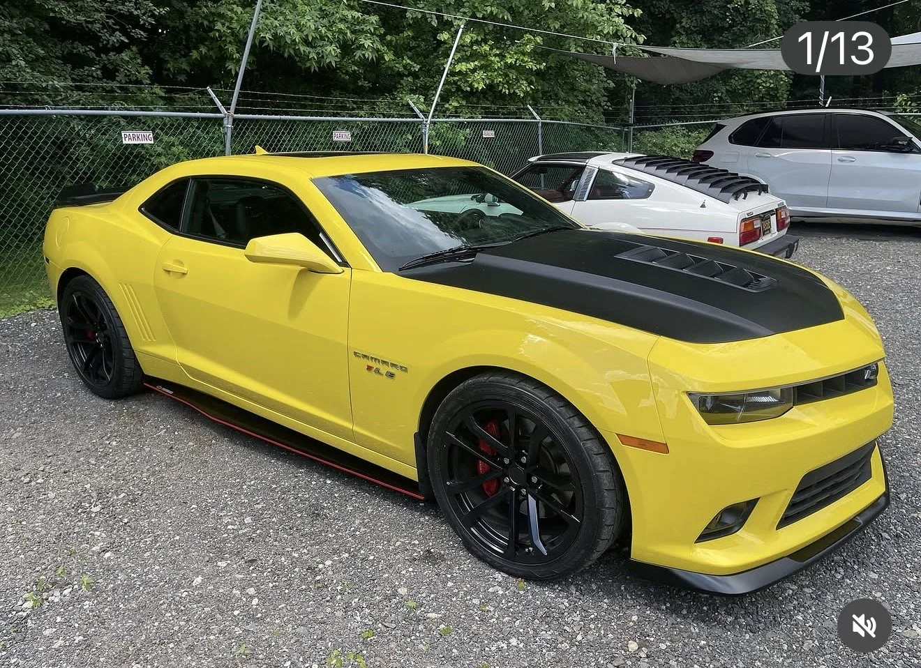 Yellow Chevrolet Camaro with black hood, black rims, and red brake calipers parked on gravel lot near a chain-link fence with trees behind, alongside a white classic car and a modern white SUV.