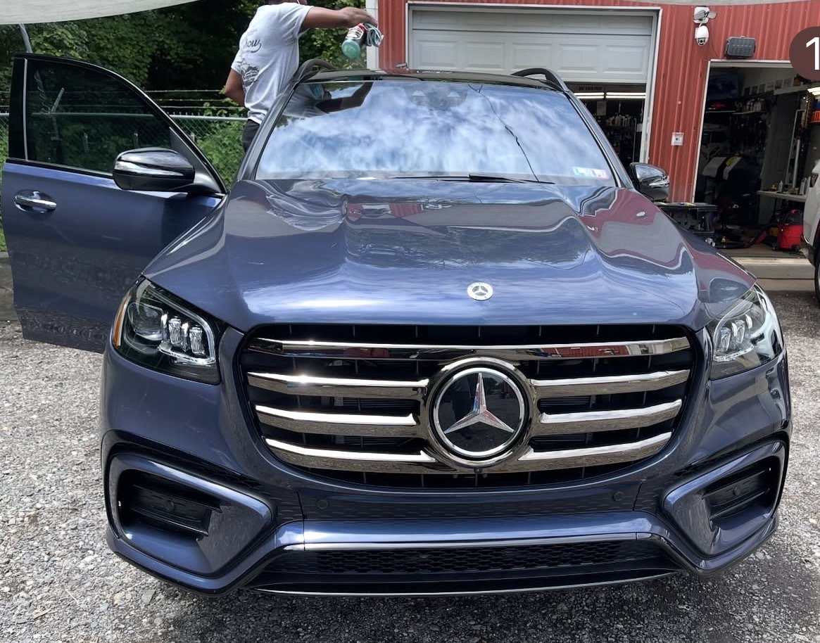 Front view of a dark gray Mercedes-Benz SUV parked outside a red garage with its driver’s side door open and a person washing the roof with a spray bottle.