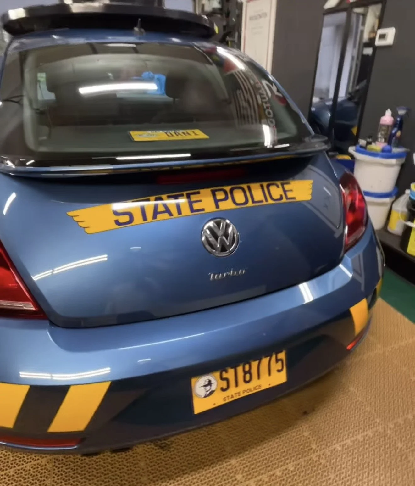 A blue Volkswagen car with a yellow and purple 'State Police' sticker on the trunk, parked indoors on a brown rug near shelves with cleaning supplies and other items. The license plate reads '5187775' and has a police emblem.