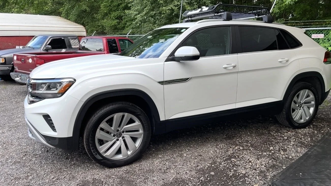 White Volkswagen SUV parked on gravel with other vehicles in the background and trees behind.