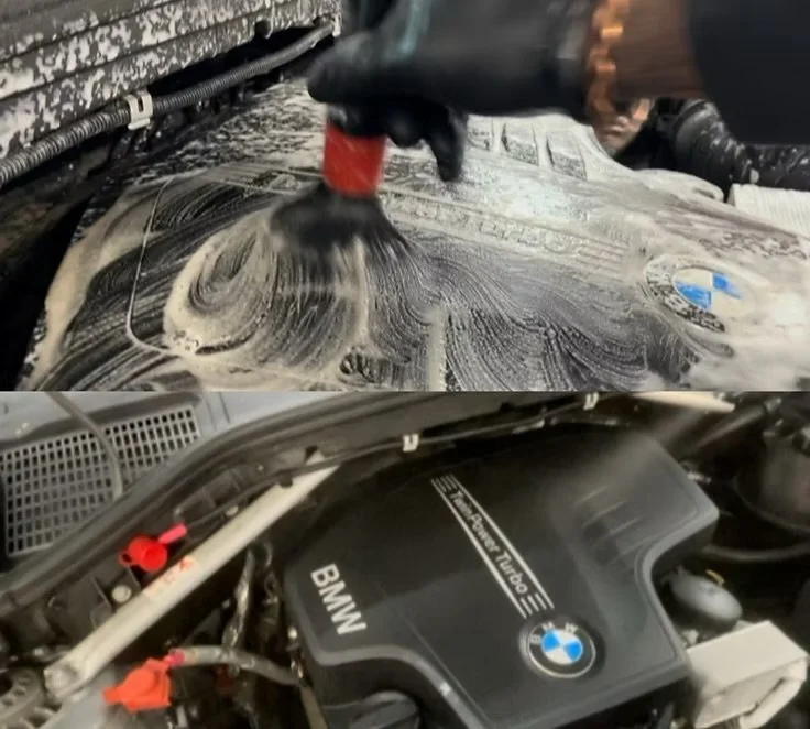 A person washing a BMW car engine with soap and water in a garage or workshop.