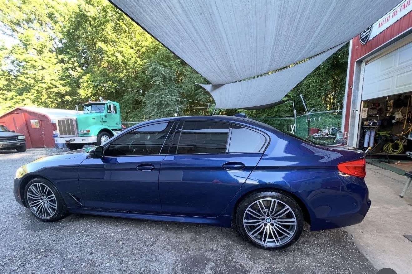 A blue sedan car parked outside a garage with two shaded sun sails overhead, a green truck in the background, and trees surrounding the area.