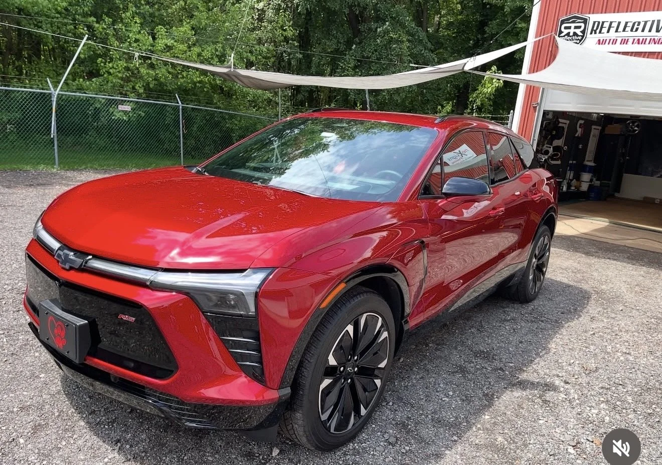 A red Chevrolet electric vehicle parked outside a service shop with a partially open garage door, surrounded by green trees and a chain-link fence.