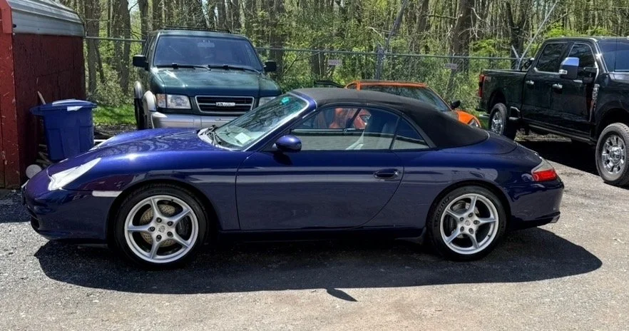 Blue convertible sports car parked on asphalt, with a black soft top, surrounded by trucks and a wooded area.