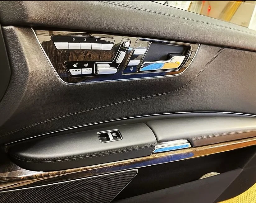 Close-up of a luxury car door interior showing leather conditioning and seat adjustment controls, heated seat buttons, window control, and wood trim.