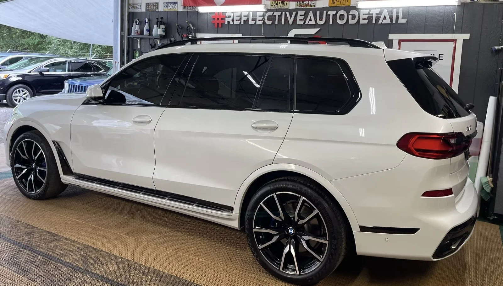 A white BMW SUV parked inside a detailing shop, with car cleaning supplies on the wall and other cars visible outside through the window.