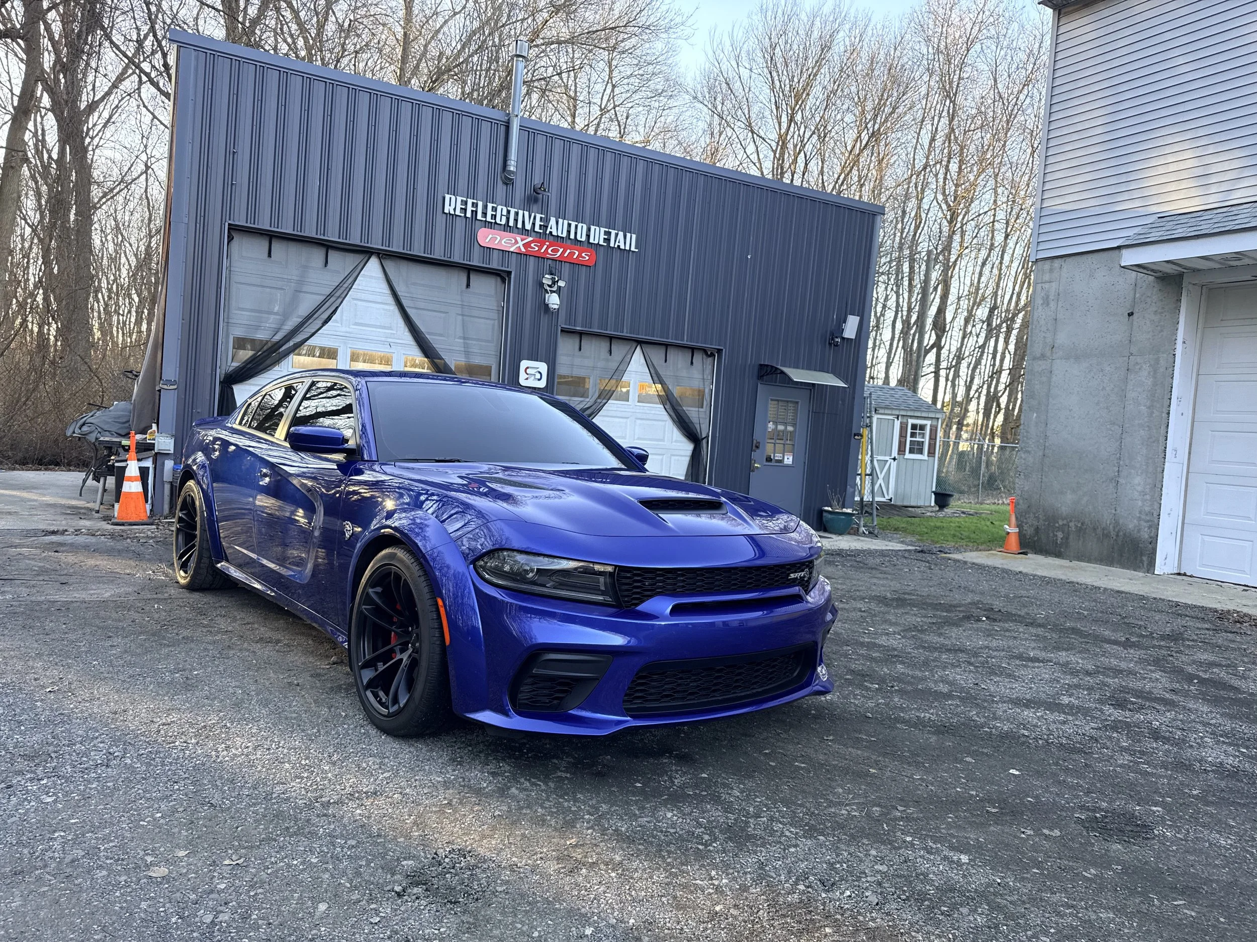 A blue sports car parked in front of a gray building with a garage door, with trees in the background and leafless branches, during the daytime.
