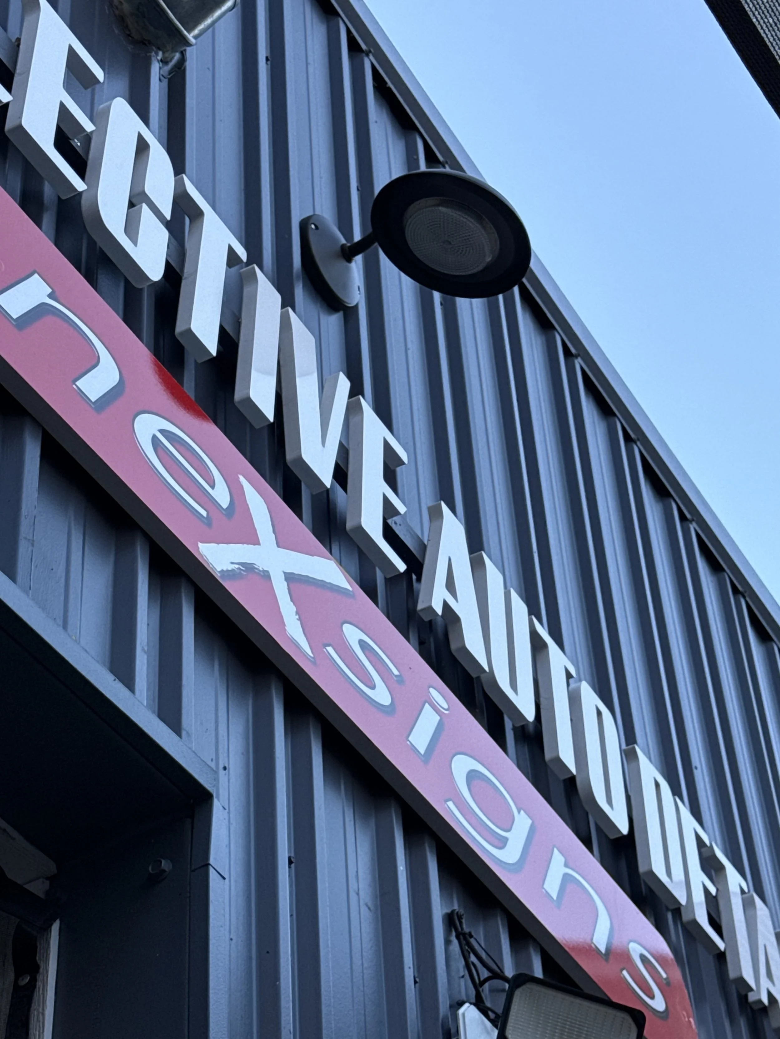 Close-up of a building exterior with a large, three-dimensional white sign spelling 'RE' vertically mounted. A black security camera is attached to the wall above the sign, and part of a pink banner with white text is visible at the bottom left corner. The building has vertical metal siding, and the sky is clear blue.