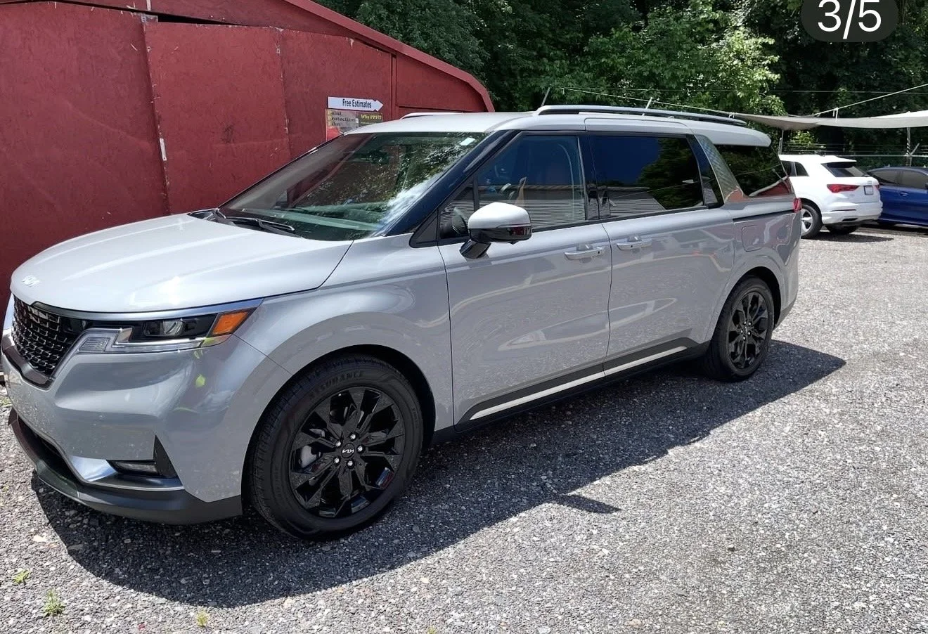 A silver minivan parked on gravel in front of a red building with trees in the background.