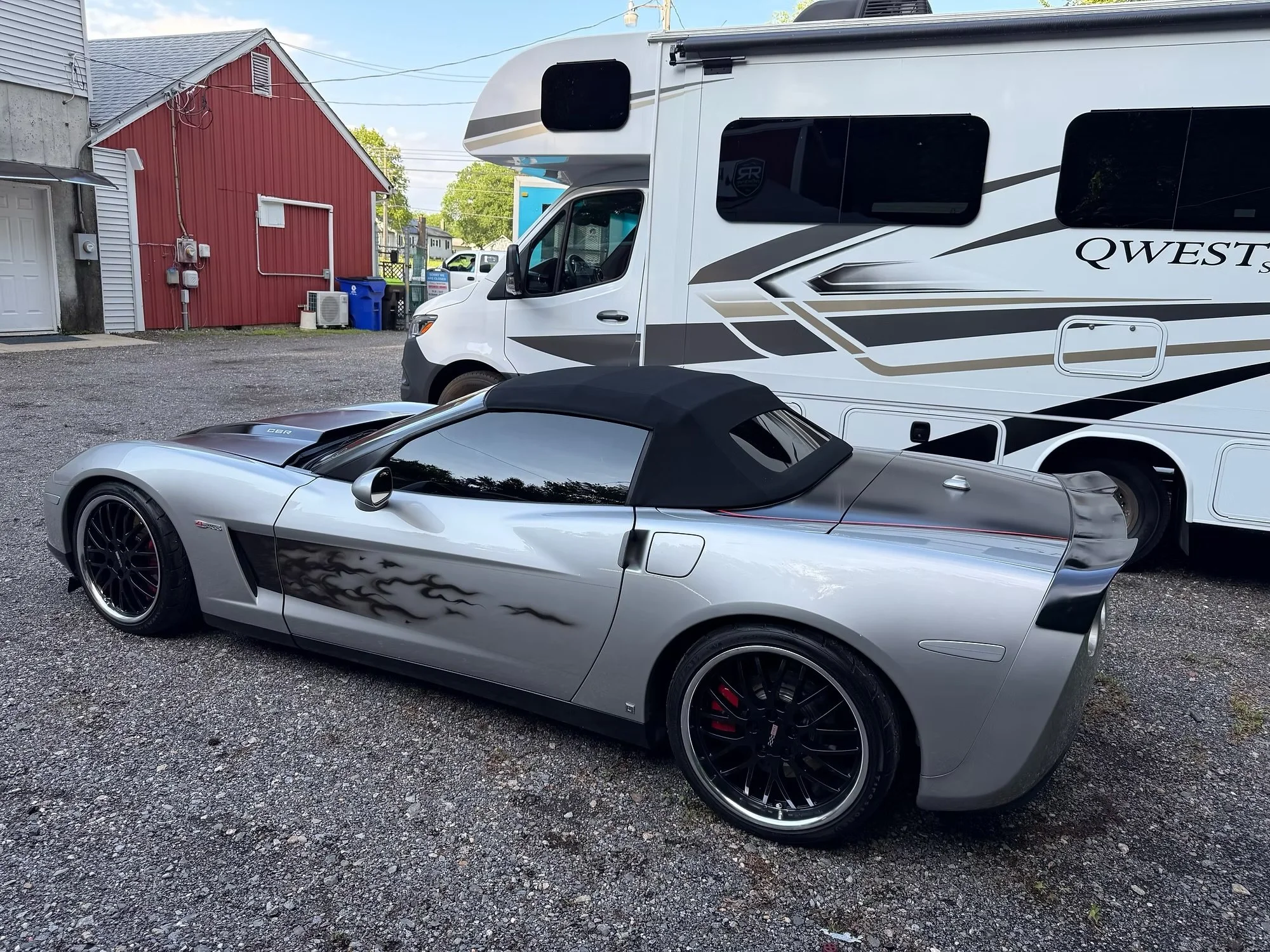 Silver convertible sports car parked next to a white RV with black windows on a gravel lot.