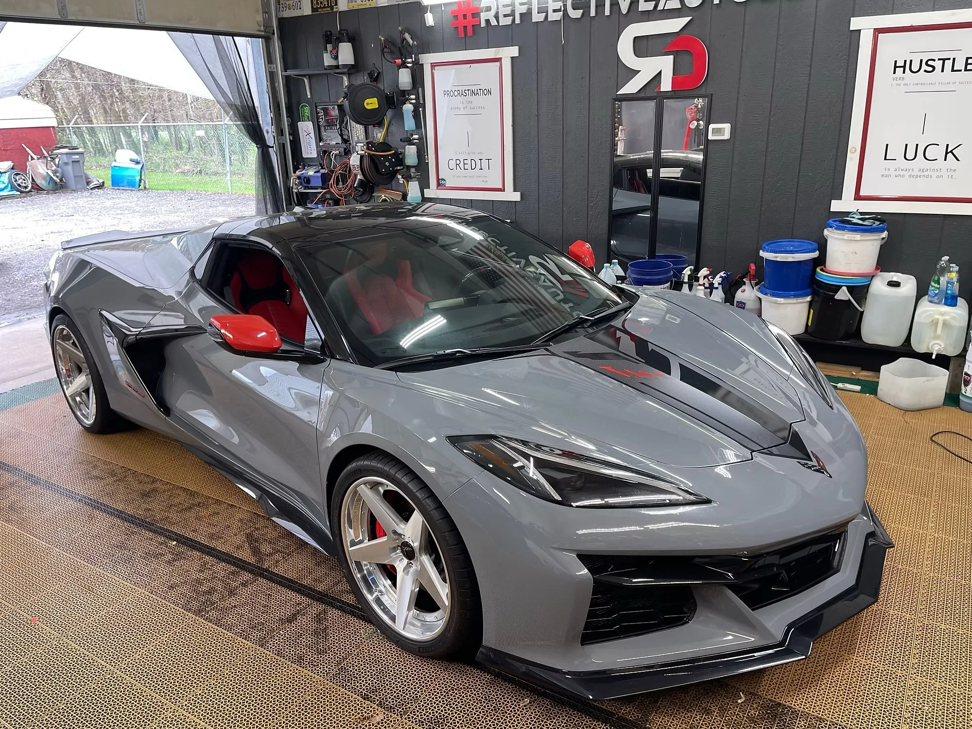 A sleek Corvette parked inside a garage or workshop with car cleaning supplies, tools, and inspirational posters on the walls.