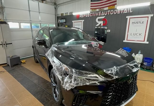 Black sedan with front-end damage and protective plastic wrap on the fender inside a garage or workshop.