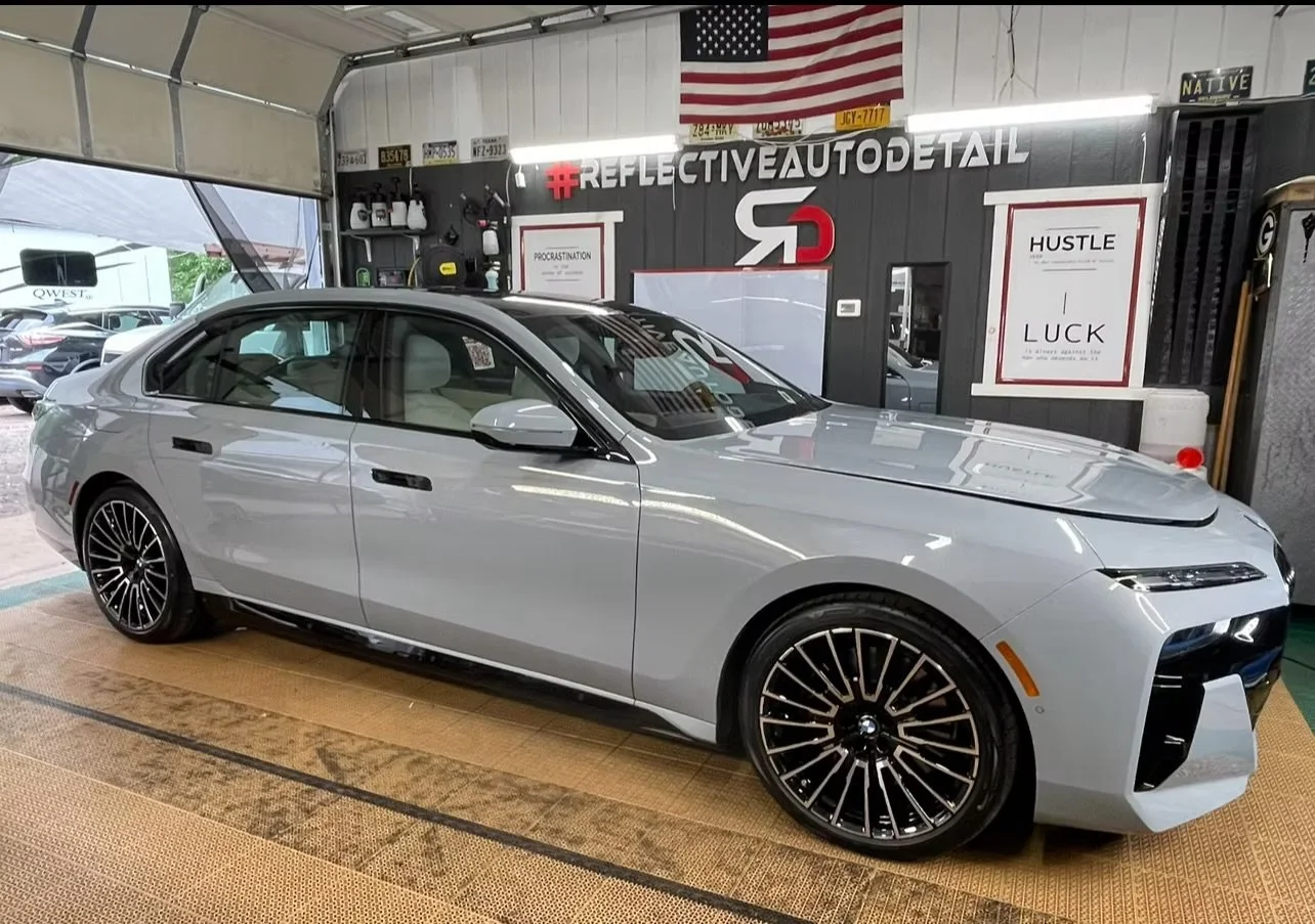 Silver sedan car inside a garage with black walls, American flag, various tools, and motivational signs on the wall, including '#REFLECTIVEAUTODETAIL' and 'HUSTLE, LUCR'.