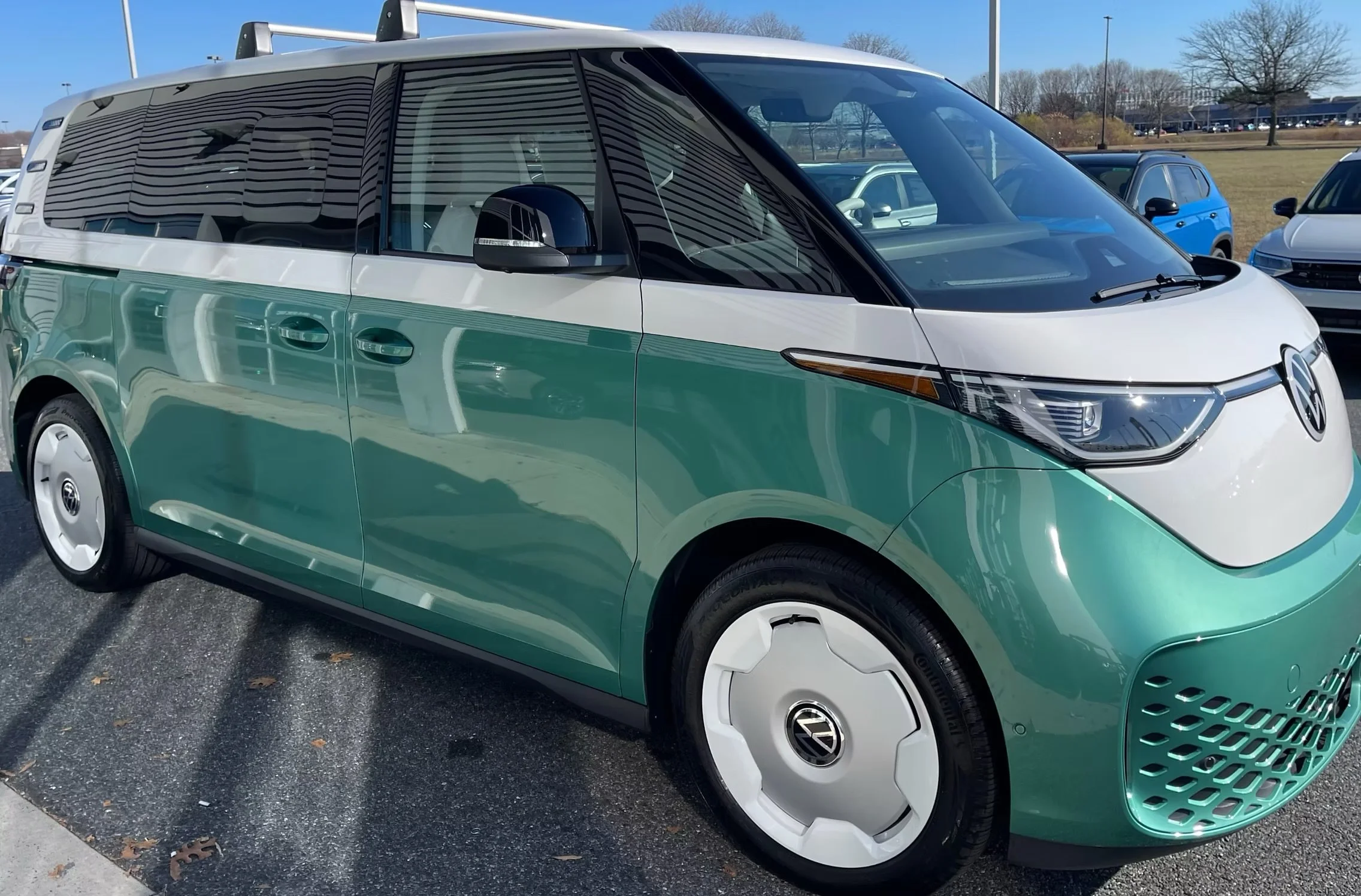 A green and white Volkswagen electric minivan parked in a parking lot on a sunny day with other cars and trees in the background.