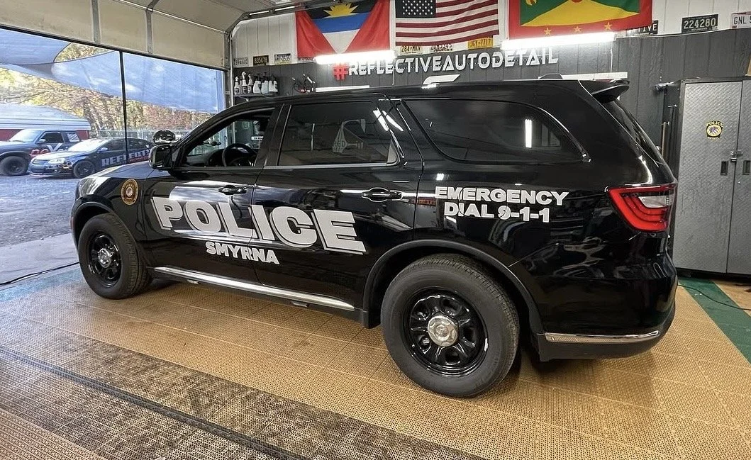 Black police SUV with 'POLICE' and 'SMYRNA' written on the side, parked indoors with flags hanging above, reflecting a well-lit interior.