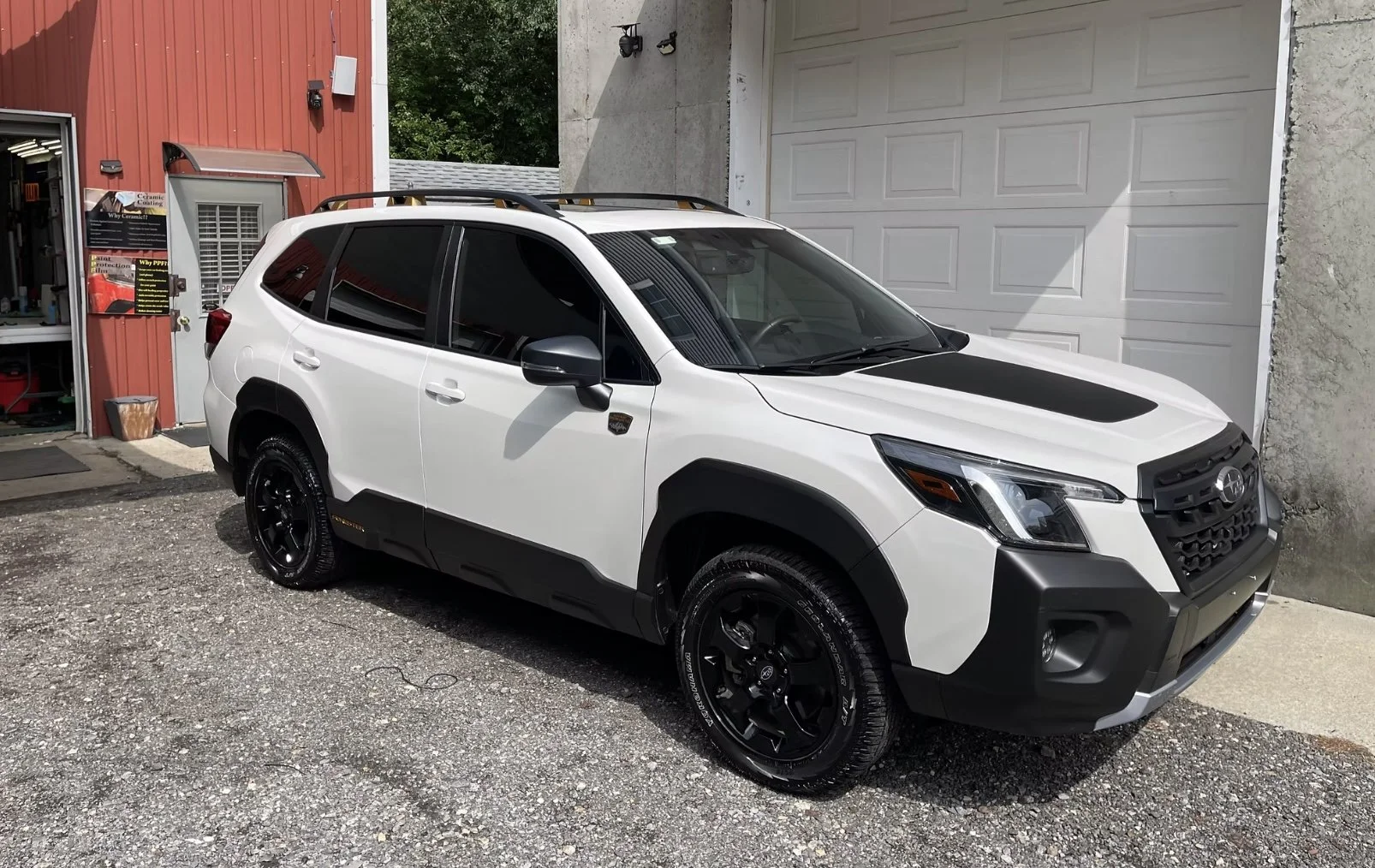 White Subaru SUV with black accents parked outside a garage, with a red building to the left and a white garage door in the background.