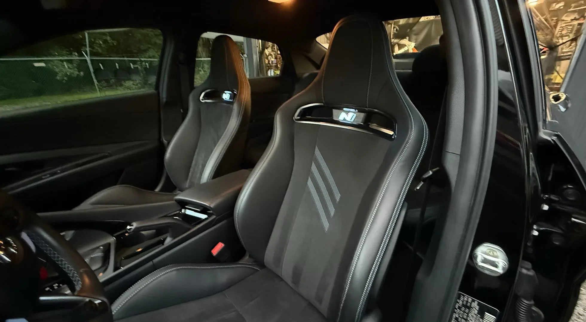The interior of a modern car showcasing two racing-style seats with a digital display on the headrests, a sleek center console, and black interior trim. Outside the windows, a fence and greenery are visible.