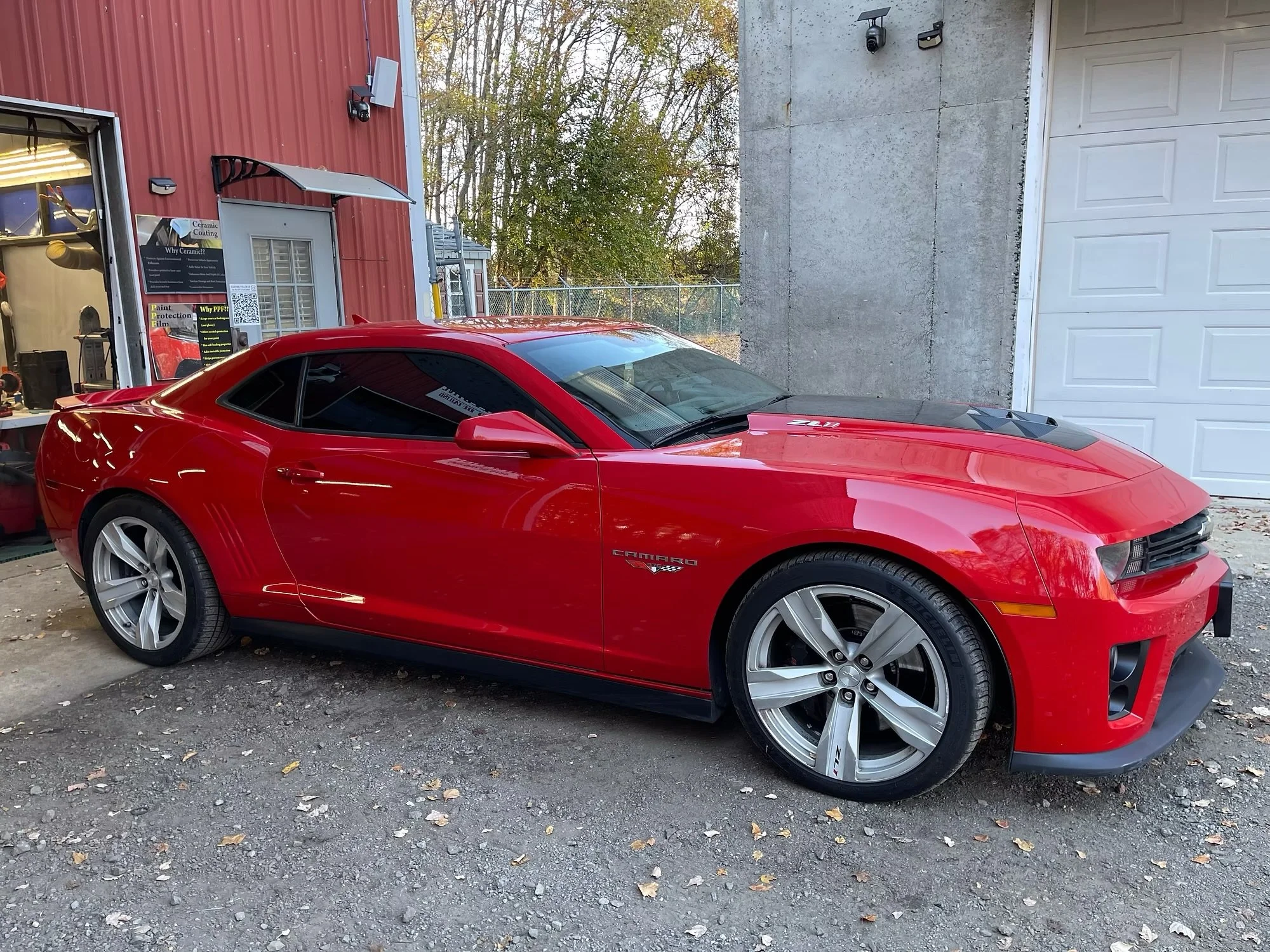 A red Chevrolet Camaro sports car is parked outside in a gravel area next to a building with red siding and a white garage door. The car has tinted windows and large silver wheels.