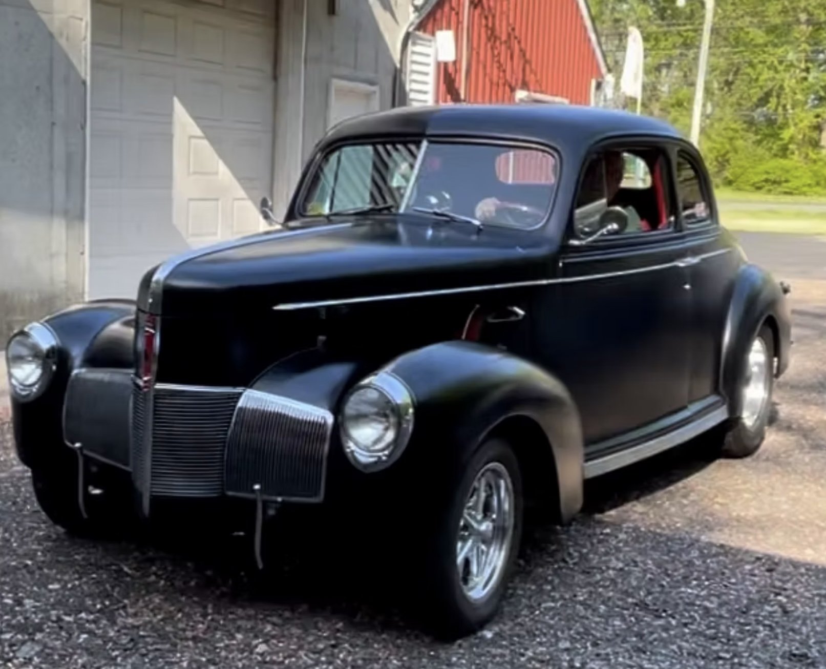 A vintage black car parked outside a garage with a white door and a red roof. The car has a rounded body, chrome details, and appears to be from the 1930s or 1940s.