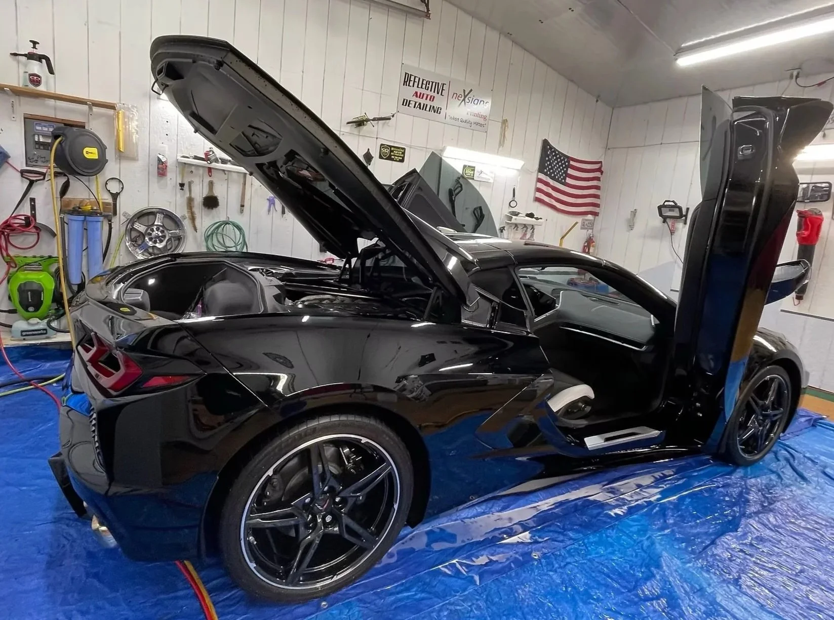 A black sports car with gullwing doors open inside a garage or detailing shop with tools on the walls and an American flag, and blue tarps on the floor.