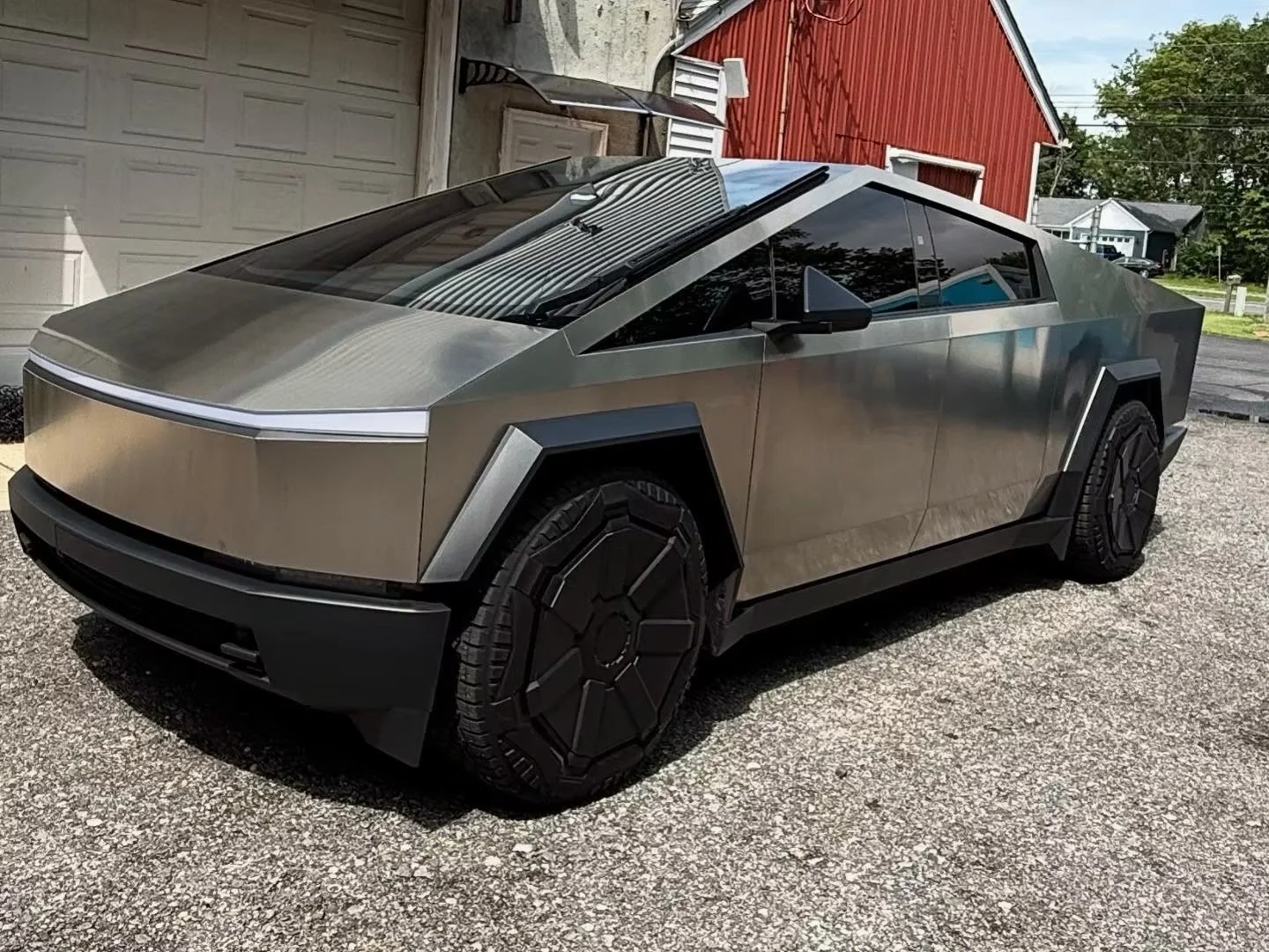 A silver CyberTruck with black wheel covers, parked on a gravel driveway in front of a garage and residential houses.