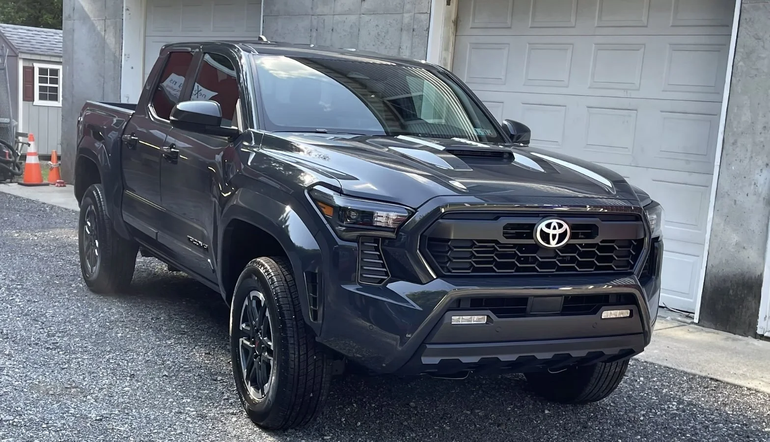 Black Toyota pickup truck parked outdoors in a gravel driveway next to a garage with a closed white door.