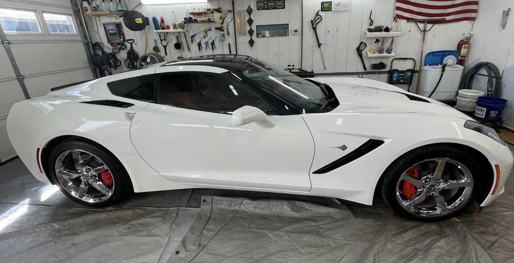 A white sports car parked inside a garage with various tools hanging on the wall and cleaning supplies in the background.