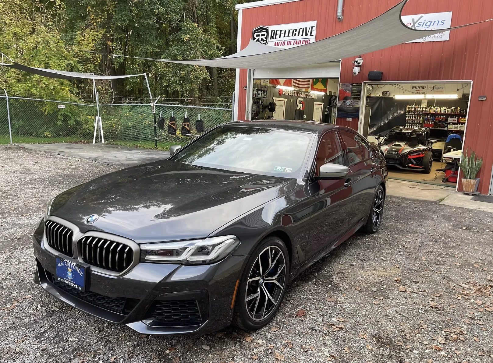 A black BMW sedan parked outside an auto detailing shop with a red and white sign, an open garage revealing a black sports car inside, and various car care products and equipment