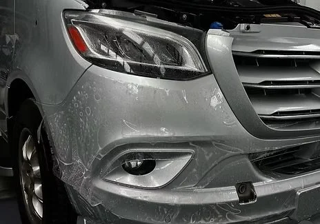 Close-up of the front right side of a gray car with visible water spray and dirt, showing the headlight, fog light, and part of the front bumper with a parking sensor.