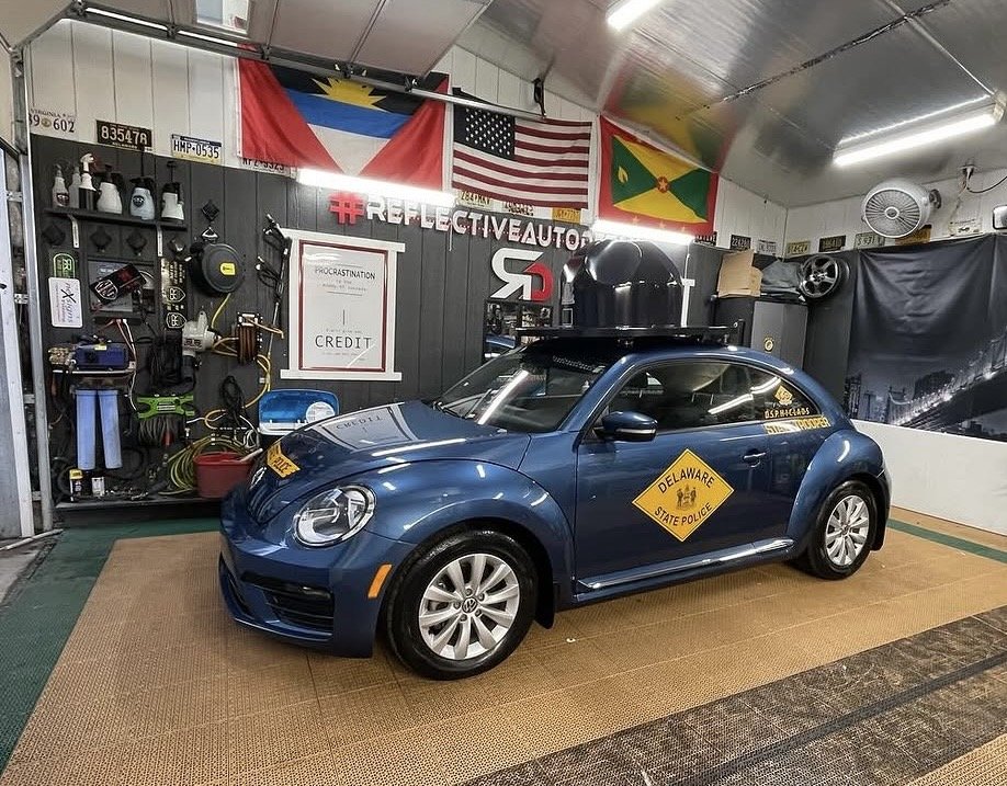 A blue Volkswagen Beetle car inside a garage with flags of the Philippines, the United States, and Guyana hanging above. The car has a Delaware State Police logo sticker on the side and a roof-mounted device. The garage has various car tools and acce